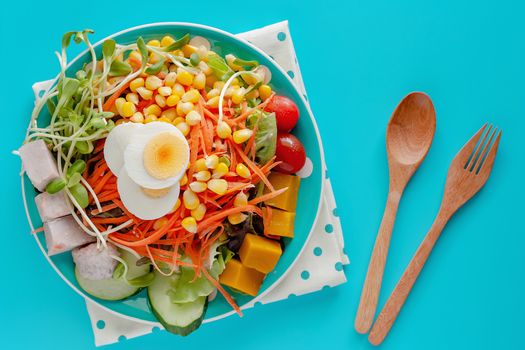 Fresh salad vegetable with boiled chicken egg, wooden spoon and fork on blue background for healthy eating and diet food concept