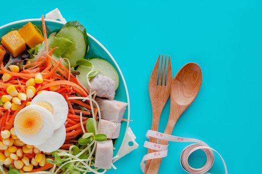 Fresh salad vegetable with boiled chicken egg, measuring tape, wooden spoon and fork on blue background for healthy eating and diet food concept