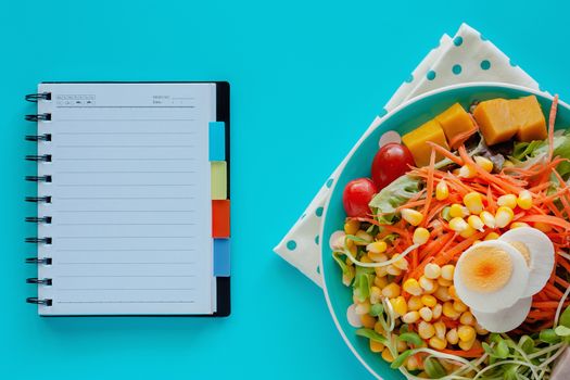 Fresh salad vegetable with boiled chicken egg and blank spiral notebook on blue background for healthy eating and diet food concept