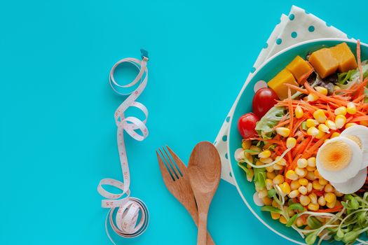 Fresh salad vegetable with boiled chicken egg, measuring tape, wooden spoon and fork on blue background for healthy eating and diet food concept