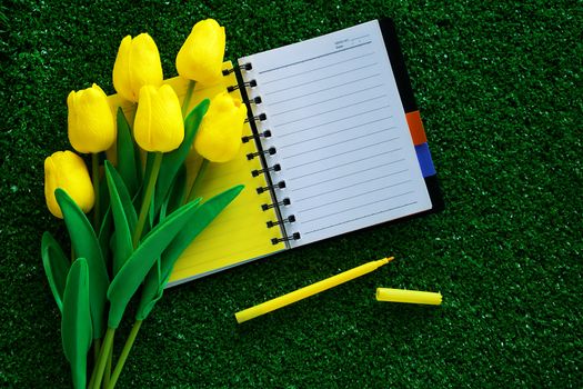 Blank notebook with artificial yellow tulips and a pen on green grass background for workspace, nature decoration and springtime concept