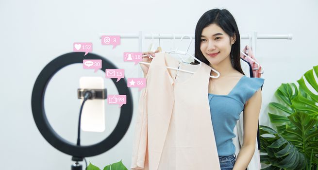 Asian woman blogger live streaming for review dress fashion accessories online on social media via mobile phone with notification interactions icons from friend in social network with like, message