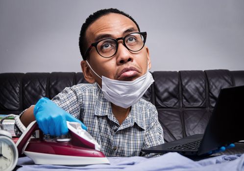 man with boring face working with very busy business and housework part, ironing cloth and working with laptop during self isolation to avoid spreading illness transmission of COVID-19 Coronavirus