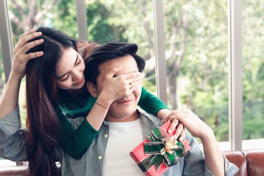 Beautiful Woman is Surprising With Gift Box While Closing Her Boyfriend Eyes at Their Home. Handsome Man Having Happy Surprise While Waiting Girlfriend Giving Presents Box. Couple Valentine Concept