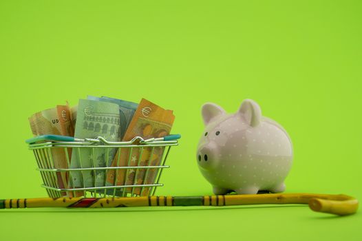 Saving for old-age and retirement concept with shopping basket filled with cash banknotes, walking stick and piggy bank over a green background with copy space
