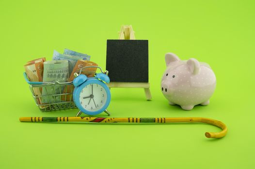 Saving for old-age and retirement concept with shopping basket filled with cash banknotes, alarm clock, walking stick, chalkboard and piggy bank over a green background with copy space