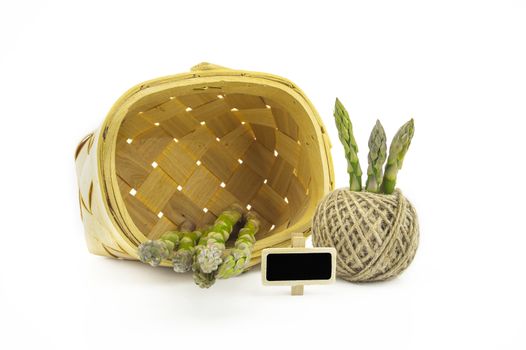 Bundle of fresh green asparagus, rustic woven basket and ball of natural hemp twine alongside a small slate or signboard isolated on white background, selective focus in a low angle view
