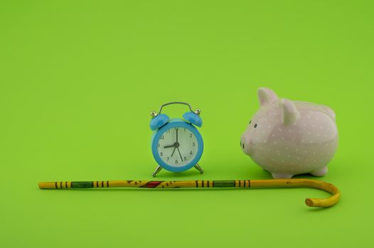 Saving for old-age and retirement concept with alarm clock, walking stick and piggy bank over a green background with copy space
