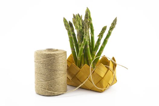 Bundle of fresh green asparagus in a rustic woven basket near ball of natural hemp twine isolated on white background, selective focus in a low angle view