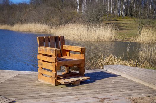 Rustic hand crafted chair made from wood pallets on wooden deck overlooking a river or lake with reeds in spring sunshine