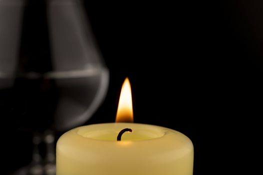 Single burning yellow candle in front of a wineglass against dark background