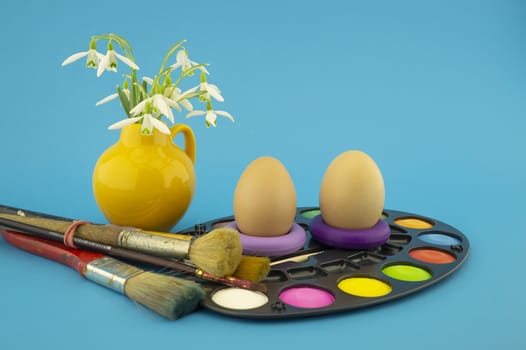 Easter egg decoration concept with paint brushes and set of watercolor paints in pallet, next to a yellow jug with fresh white spring flowers. Studio still life on blue background