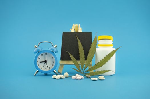 Medicinal marijuana concept with fresh green leaf, pharmaceutical bottle and pills, alarm clock and small chalk board over a blue background with copy space