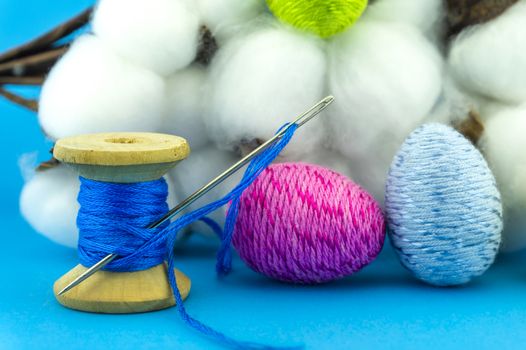Handcrafted Easter decorations from natural cotton with a cotton bolls off the plant behind a reel of blue thread with needle and creative colorful eggs made from yarn over a blue background