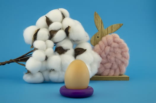 Natural cotton fluffy Easter bunny decoration with a cotton boll of the plant and hens egg on a purple ring over a colorful blue background for a holiday card design