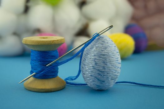 Handcrafted Easter decorations from natural cotton with a cotton bolls off the plant behind a reel of blue thread with needle and creative colorful eggs made from yarn over a blue background