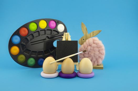 Colorful creative Easter still life with handicrafts featuring three eggs on purple rings, a natural fluffy cotton or wool rabbit and multicolored artist palette with paint and brush over blue