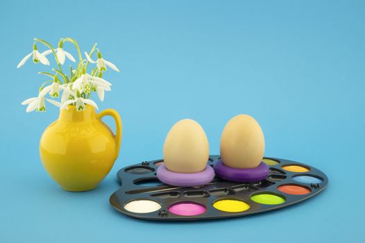 Easter egg decoration concept with set of watercolor paints in pallet, next to a yellow jug with fresh white spring flowers. Studio still life on blue background