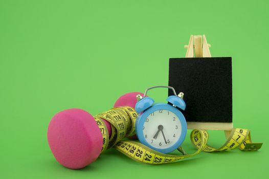 Starting healthy lifestyle concept. Still life with pink dumbbell, measuring tape, chalkboard and alarm clock on green background