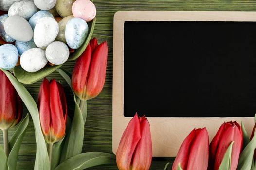 Easter background. Red spring tulips, painted eggs and blank school slate for text on vintage wooden board