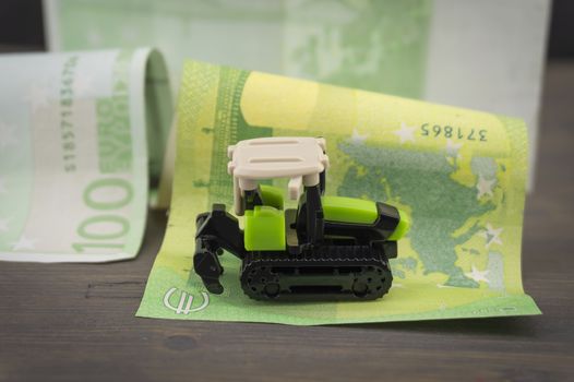 Small plastic toy heavy duty digger or tractor with caterpillar treads on 100 Euro notes on a wooden table, payments to european union farmers conceptual image