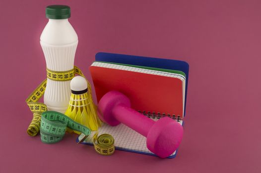 Sport weight loss and fitness concept with a shuttlecock, dumbbell weight and tape measure wound round a white plastic bottle with a slim line shape, open notebook with pen on a bright pink background