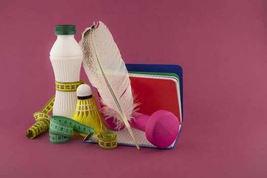 Sport weight loss and fitness concept with a shuttlecock, dumbbell weight and tape measure wound round a white plastic bottle with a slim line shape, open notebook with quill pen on a pink background