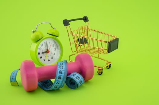 Going shopping for sports diet concept still life with dumbbells, measuring tape, alarm clock and wire shopping cart on green background