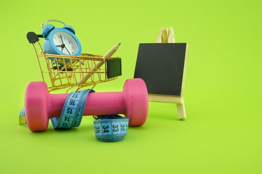 Going shopping for sports diet concept still life with dumbbells, measuring tape, blank blackboard, alarm clock and wire shopping cart on green background