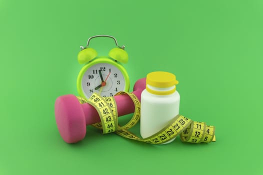 Starting healthy lifestyle concept. Still life with pink dumbbell, measuring tape and alarm clock on green background