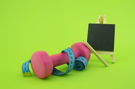 Starting healthy lifestyle concept. Still life with pink dumbbell, measuring tape, chalkboard and pencil on green background