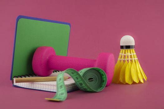 Sport weight loss and fitness concept with a shuttlecock, dumbbell weight and tape measure alongside an open notebook with pen on a bright pink background