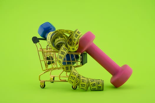 Going shopping for sports diet concept still life with dumbbells, measuring tape and wire shopping cart on green background