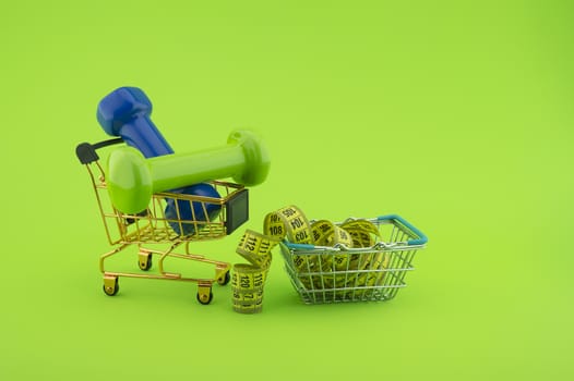 Going shopping for sports diet concept still life with dumbbells, measuring tape and wire shopping basket and cart on green background