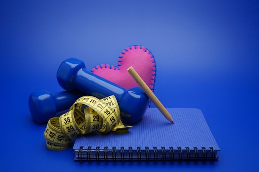 Fitness and weight loss concept with dumbbells, measuring tape, heart and new notebook on blue background