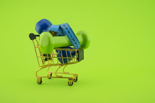 Going shopping for sports diet concept still life with dumbbells, measuring tape and wire shopping cart on green background
