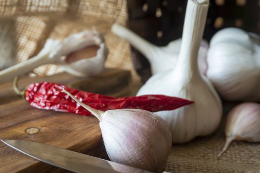 Garlic cloves, bulbs and red chilli pepper in close-up on sack cloth with kitchen knife and wooden cutting board