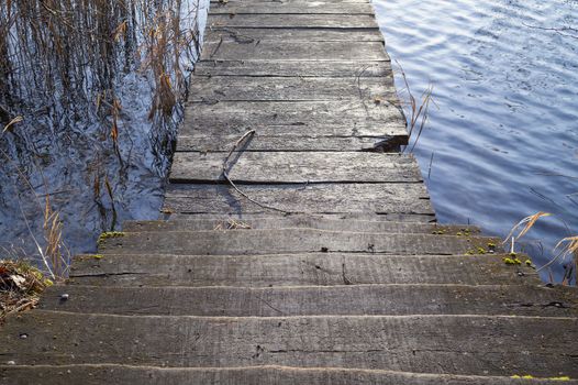 Old rustic wooden steps leading to a jetty over the calm water of a lake with reeds looking down from the top