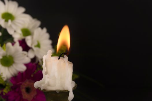 Short white candle burning on the floor in close-up among flowers against dark background.