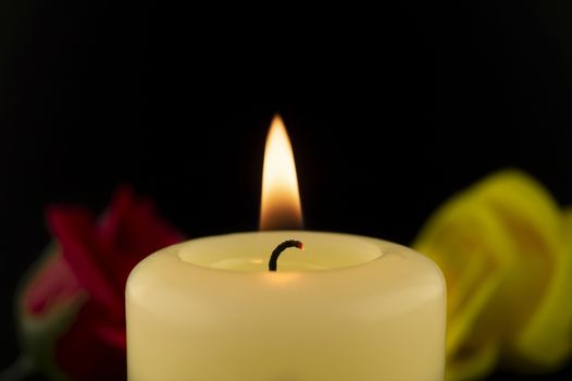 Single burning yellow candle next to red and yellow roses against dark background