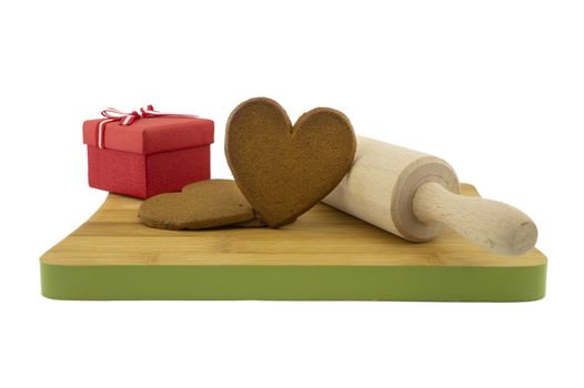 Heart-shaped cookies gift preparation concept with brown biscuits next to a gift box and rolling pin on wooden cutting board isolated on white background