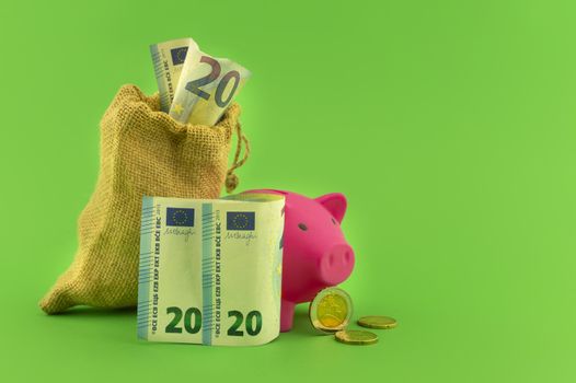 Reaping the rewards of financial success with a cute pink piggy bank, sack of money with a twenty Euro bill in the slot over a green background