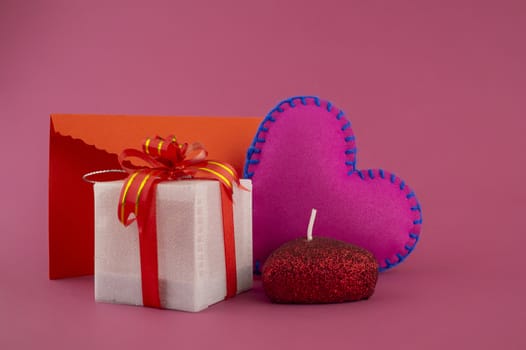 Romantic still life with hand sewn textile pink heart, candle, decorative gifts and red envelope on pink background with copy space