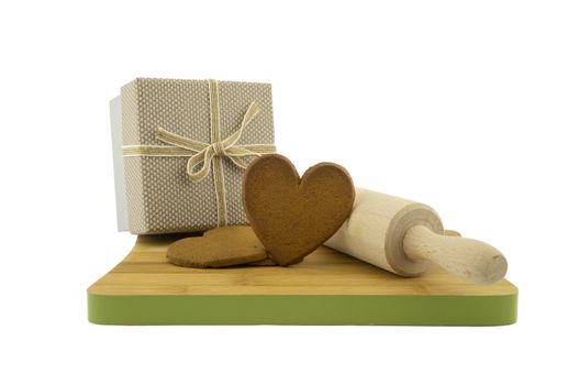Heart-shaped cookies gift preparation concept with brown biscuits next to a gift box and rolling pin on wooden cutting board isolated on white background