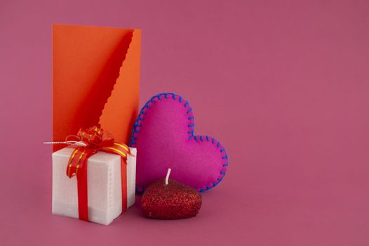 Romantic still life with hand sewn textile pink heart, candle, decorative gifts and red envelope on pink background with copy space
