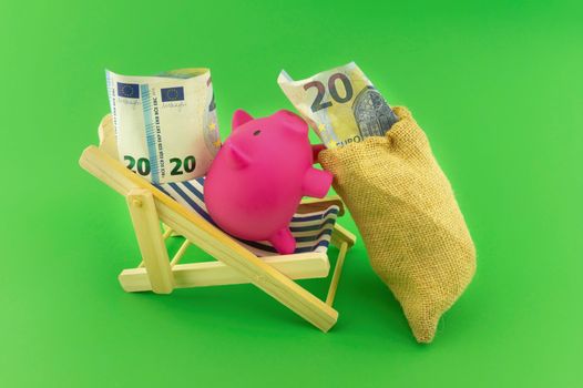 Reaping the rewards of financial success with a cute pink piggy bank relaxing on a deck chair alongside a sack of money with a twenty Euro bill in the slot over a green background