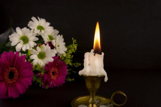 Short white candle burning on the floor in close-up among flowers against dark background.