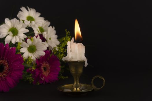 Short white candle burning on the floor in close-up among flowers against dark background.