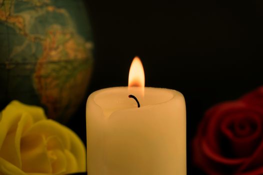 Single burning yellow candle in front of a globe next to red and yellow roses against dark background, concept of global disasters and deaths
