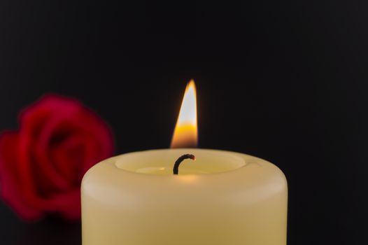 Single burning yellow candle next to red rose against dark background